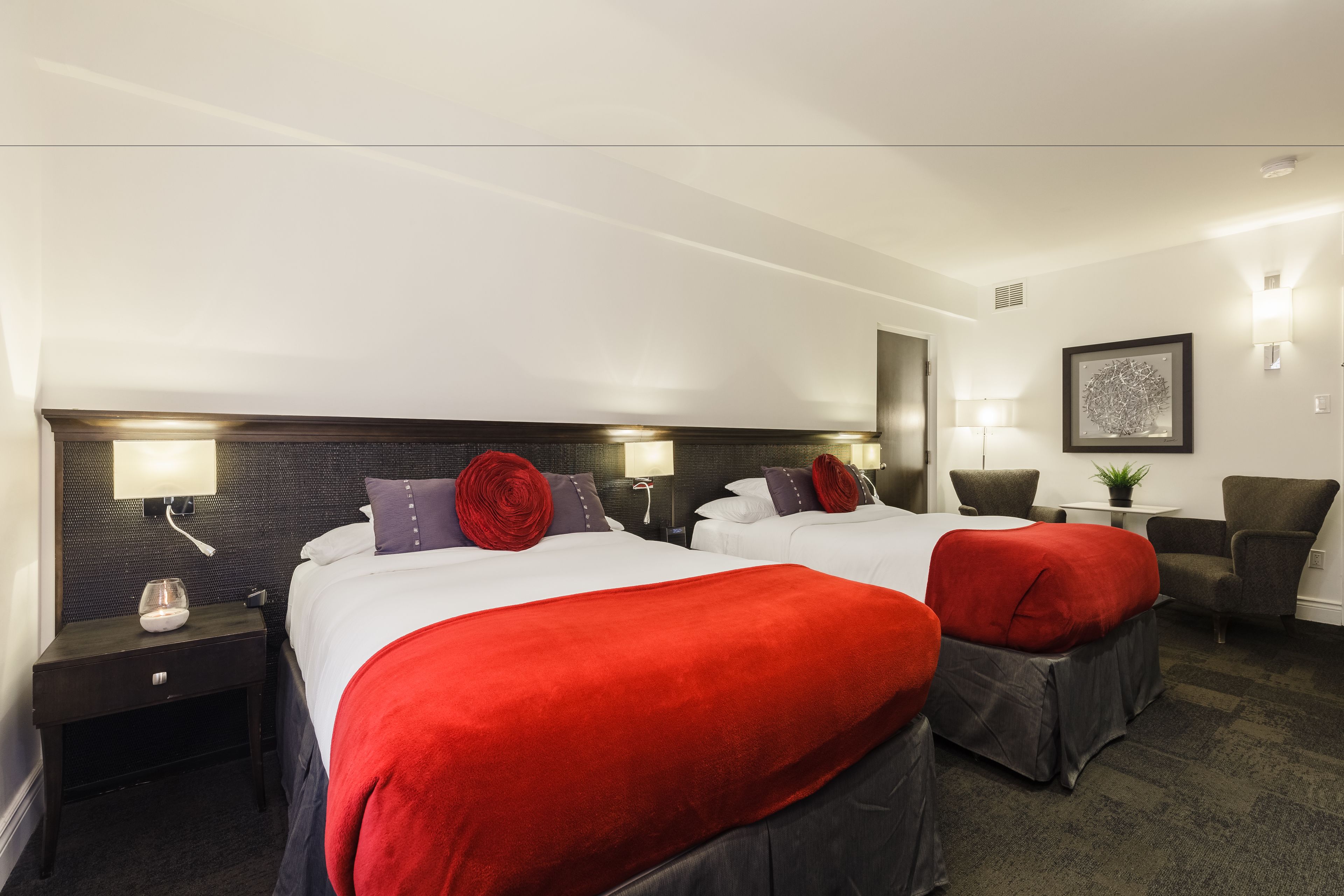 Quarto de hotel com duas camas, cada uma com roupa de cama vermelha e branca, cabeceira escura, abajures, uma mesinha com uma planta e uma cadeira.