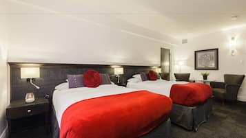 Quarto de hotel com duas camas, cada uma com roupa de cama vermelha e branca, cabeceira escura, abajures, uma mesinha com uma planta e uma cadeira.
