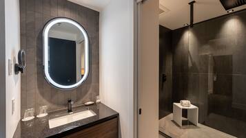 Un baño moderno con pared de azulejos oscuros, un espejo redondo con luz, un lavabo y una cabina de ducha con mampara de vidrio.