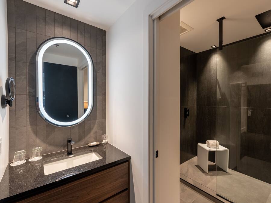 Un baño moderno con pared de azulejos oscuros, un espejo redondo con luz, un lavabo y una cabina de ducha con mampara de vidrio.