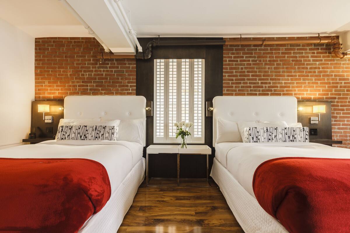 A hotel room with two beds, a brick wall, a window with blinds, and a small table with a vase of flowers.