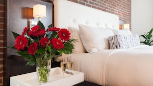 A hotel room with a brick wall, a white bed with multiple pillows, a bedside table with a lamp, and a tray with flowers and glasses of champagne.