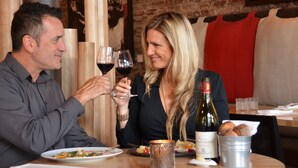 A man and a woman sitting at a table with wine glasses, a bottle of wine, and a bucket of bread.