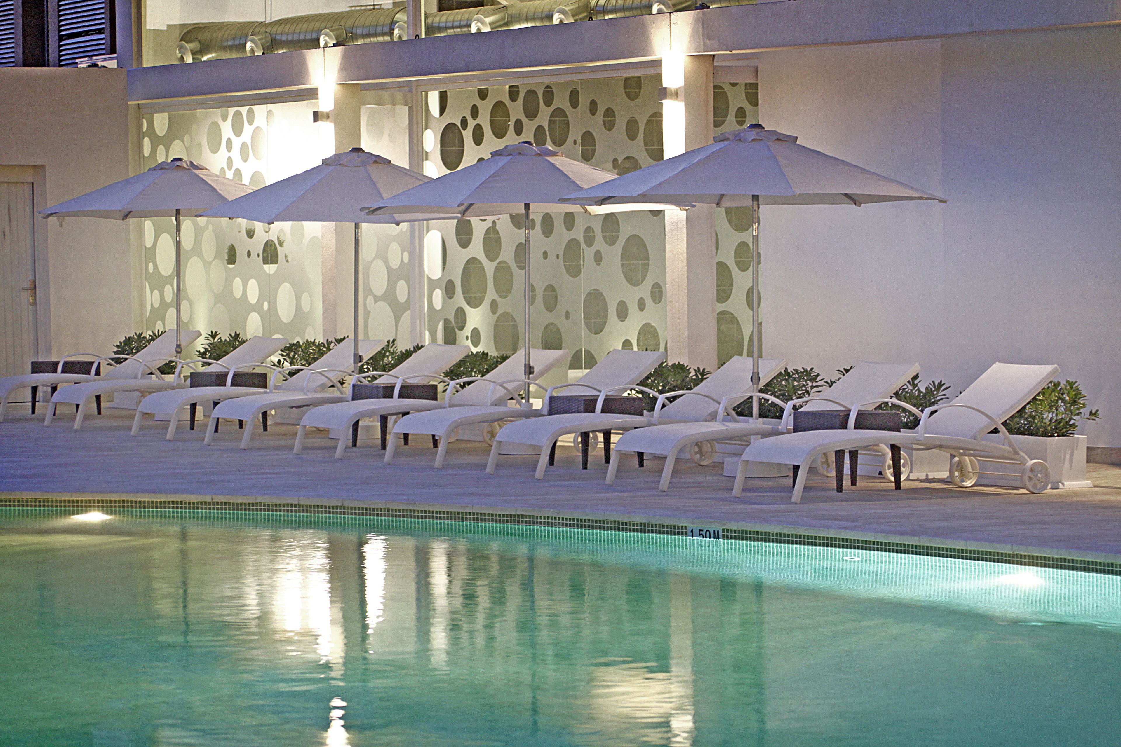 Un área junto a la piscina con sillones de descanso y sombrillas, una piscina con azulejos y una pared con diseños circulares.