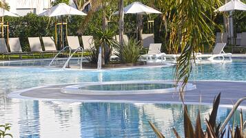 Zona de piscina con sillones de descanso y sombrillas, rodeada de palmeras y una piscina azul cristalina.