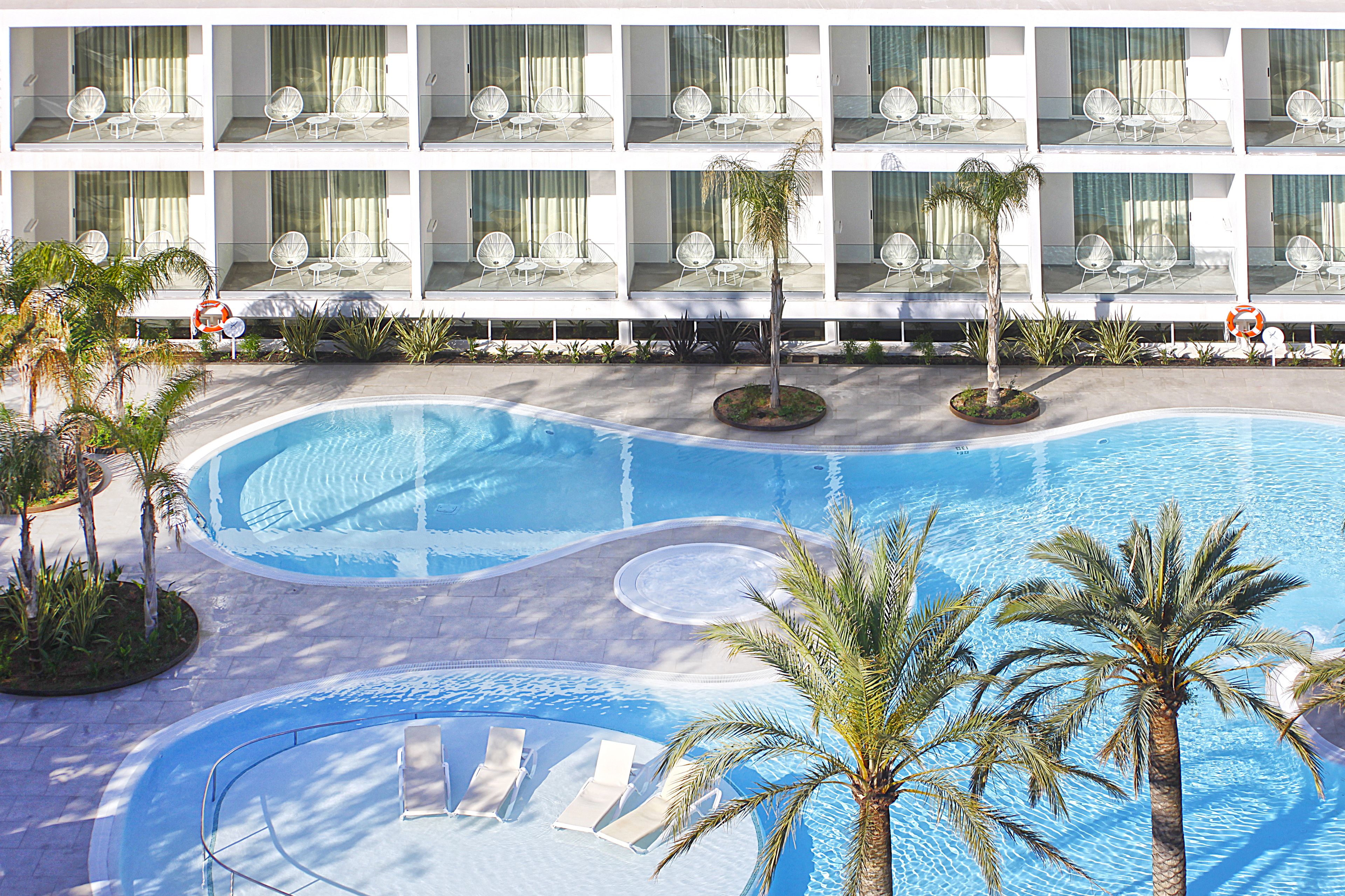 A hotel with a swimming pool, multiple balconies with chairs, and palm trees.