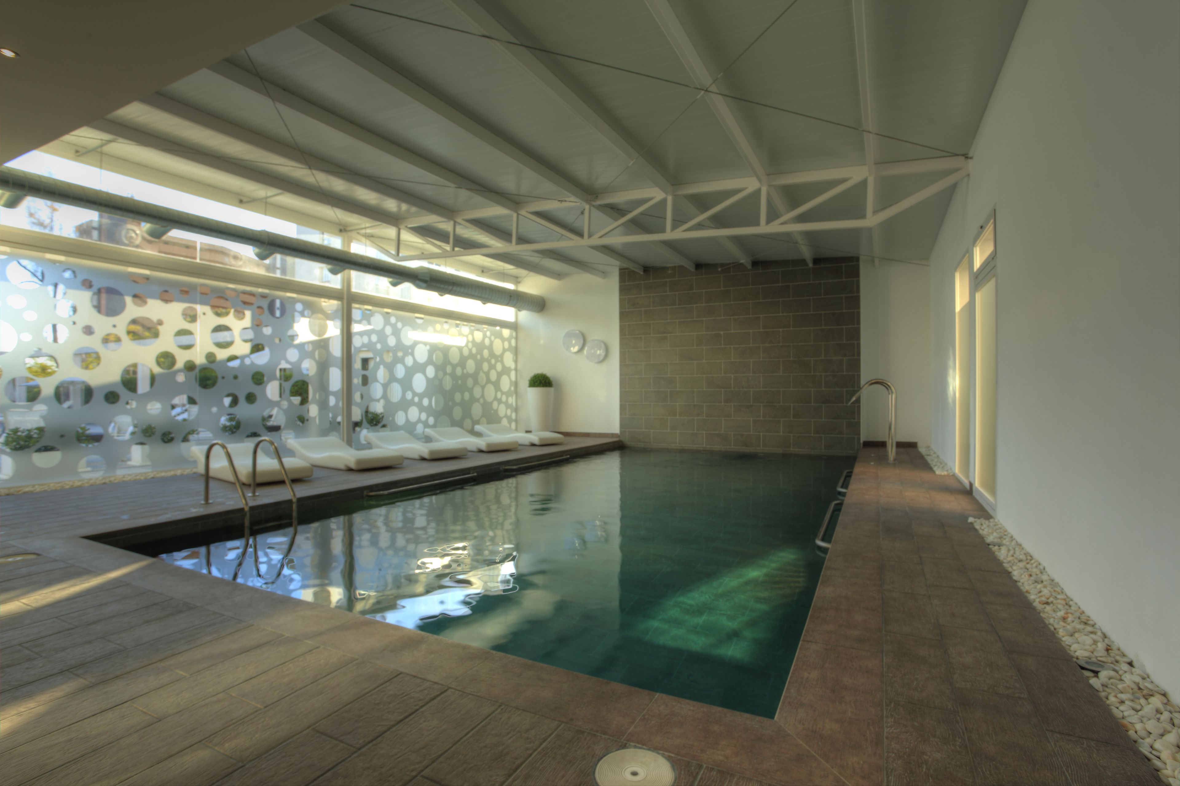 An indoor swimming pool with a wooden deck, a brick wall, and a glass wall with circular patterns.