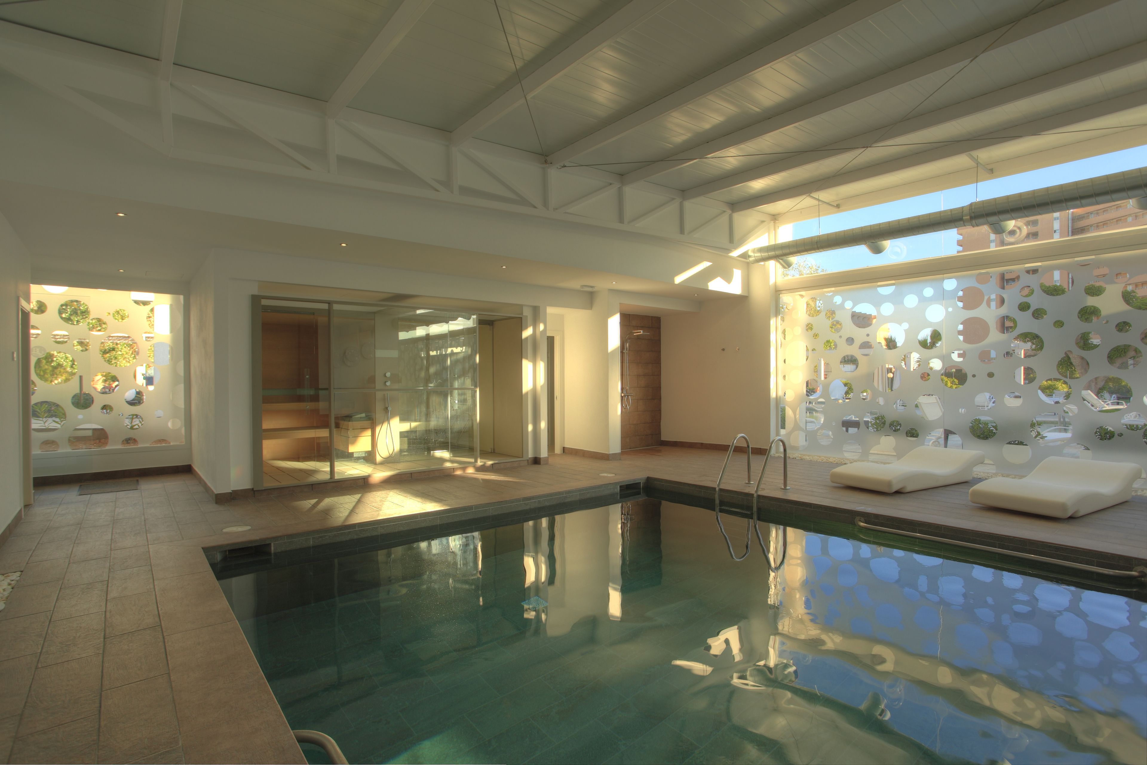 A modern indoor swimming pool with clear glass walls, a tiled floor, and a ceiling with exposed beams.