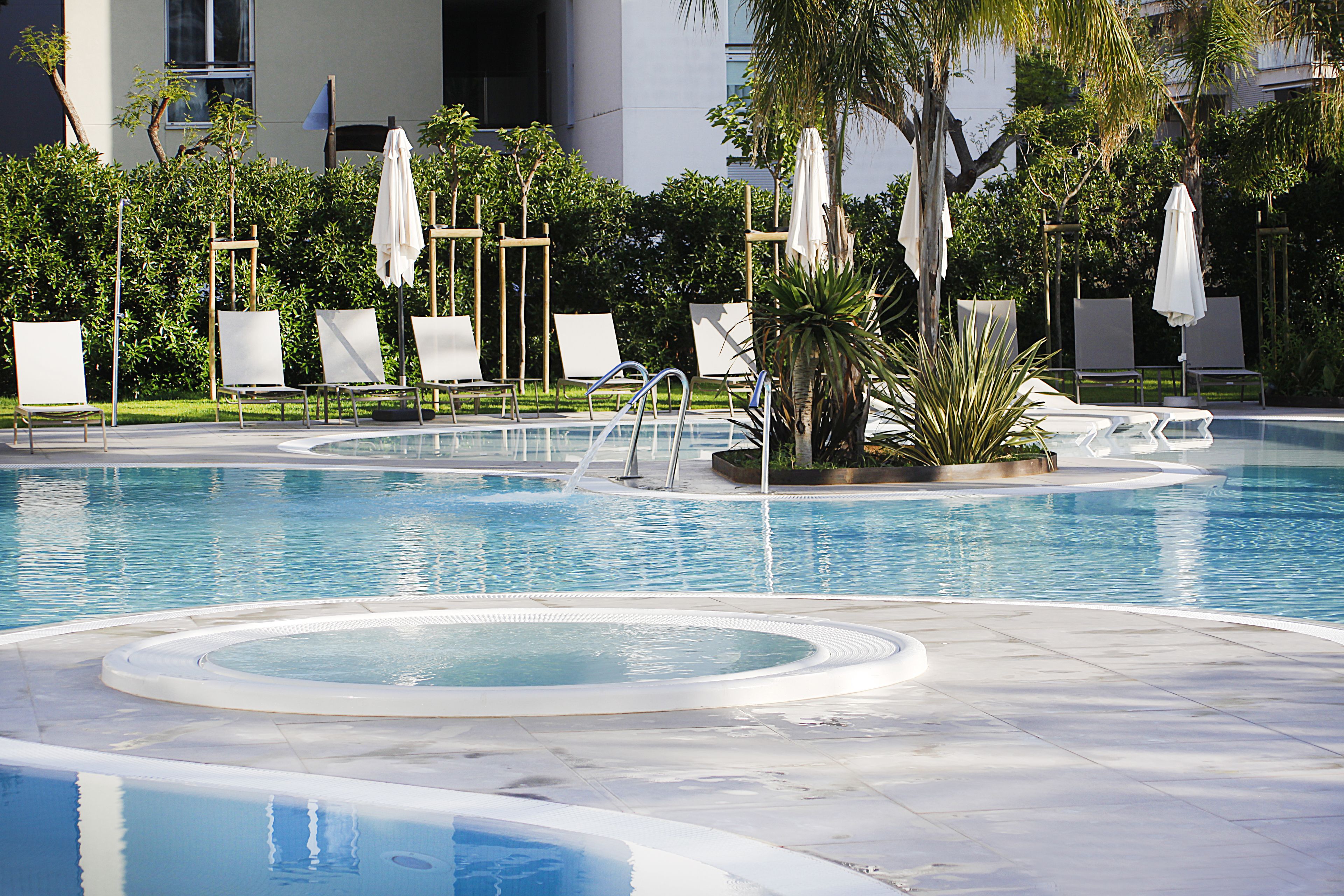 A swimming pool with multiple lounge chairs and umbrellas, surrounded by greenery and a building in the background.