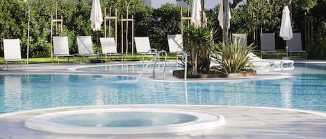A swimming pool with multiple lounge chairs and umbrellas, surrounded by greenery and a building in the background.