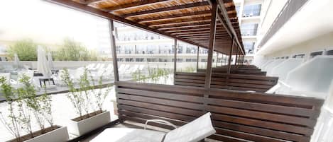 A covered outdoor seating area with white chairs and a wooden bench, overlooking a parking lot and buildings.