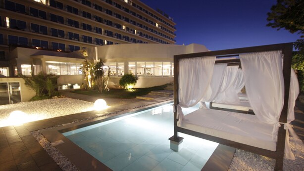 Eine Liege am Pool mit weißen Vorhängen, ein Pool mit klarem Wasser und ein Hotelgebäude im Hintergrund.