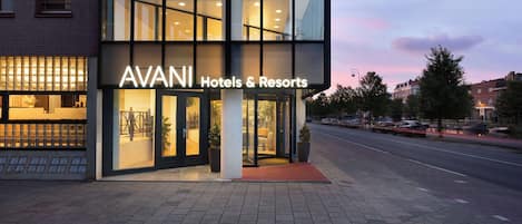 Un hotel moderno con fachada de vidrio, llamado AVANI Hotels & Resorts, que cuenta con una alfombra roja en la entrada y un área de estar al aire libre visible.