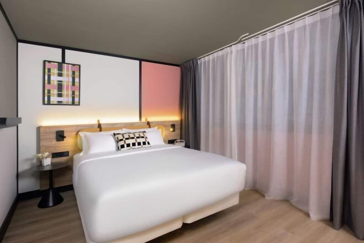 Une chambre d’hôtel moderne avec un grand lit, une table de chevet, une œuvre d’art accrochée au mur et une vue sur le coucher de soleil à travers les rideaux.