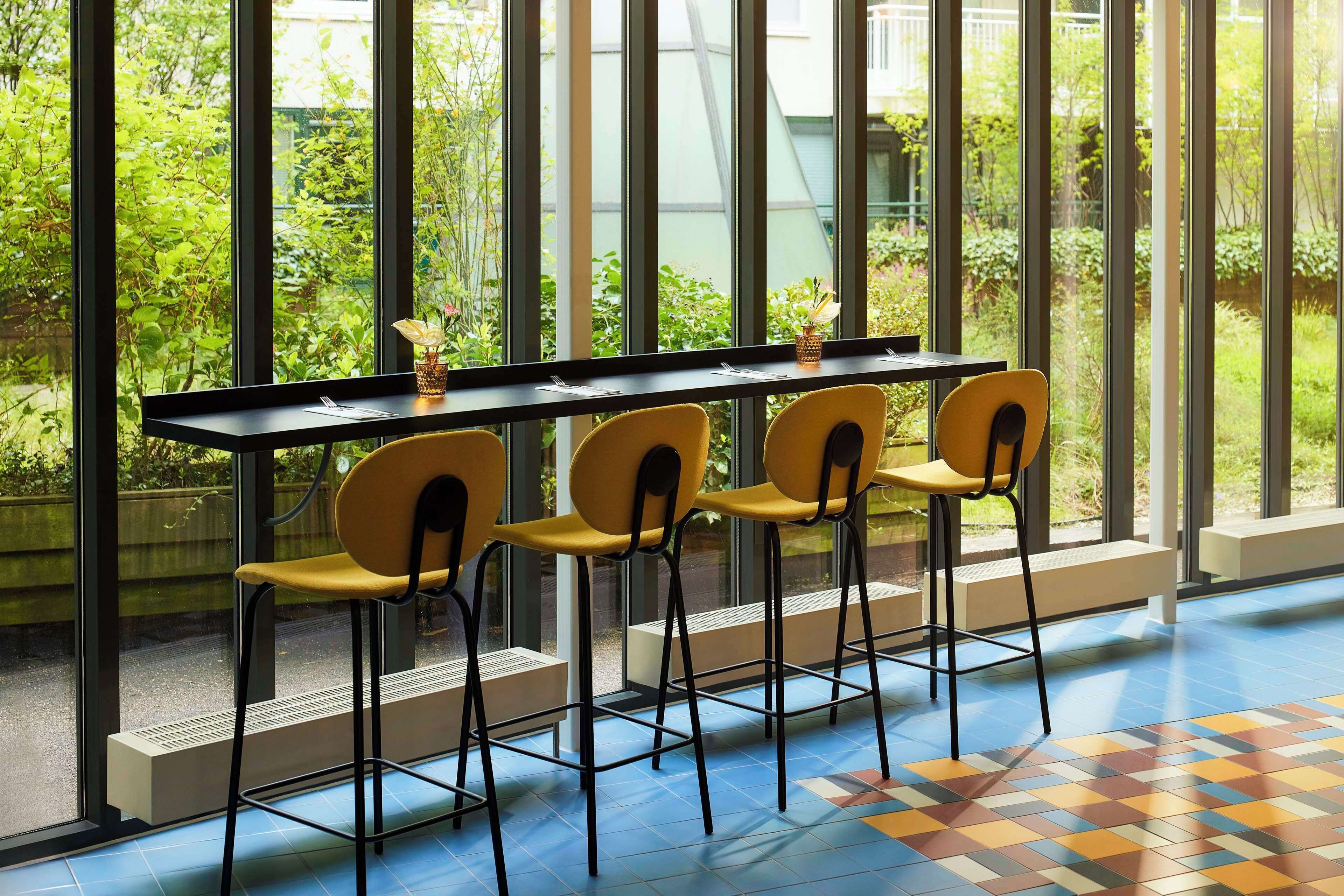 Un espace repas comprenant une longue table et quatre chaises, de grandes fenêtres donnant sur un paysage verdoyant et un sol à motifs.