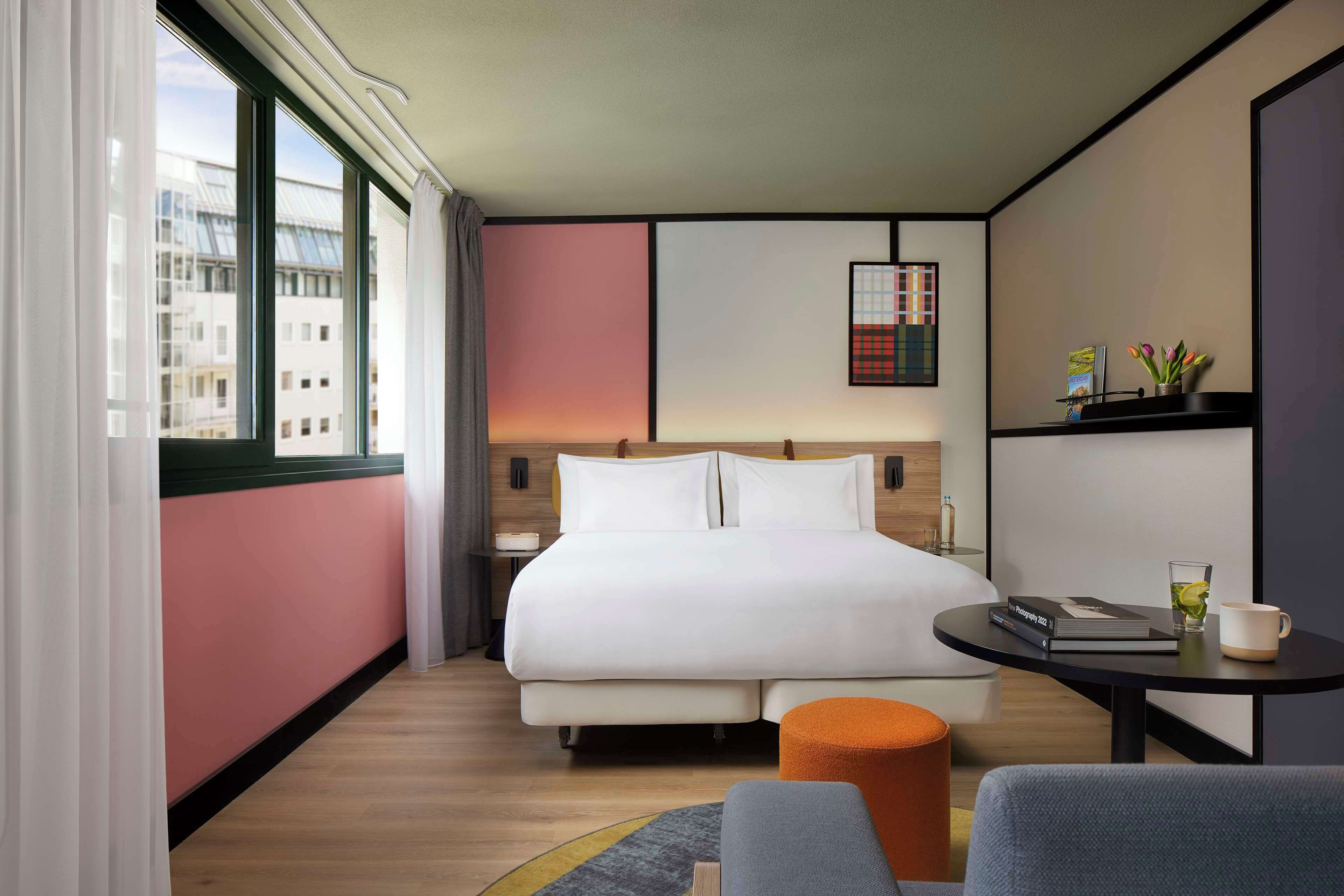 A modern hotel room with a large bed, a small round table, a chair, and a view of the city through large windows.