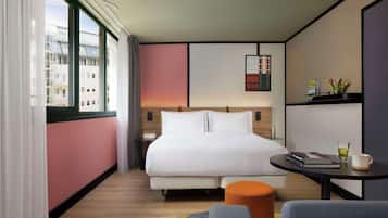 Habitación de hotel moderna con una cama grande, una mesita de noche, una pequeña mesa redonda y vistas de la ciudad a través de amplias ventanas.