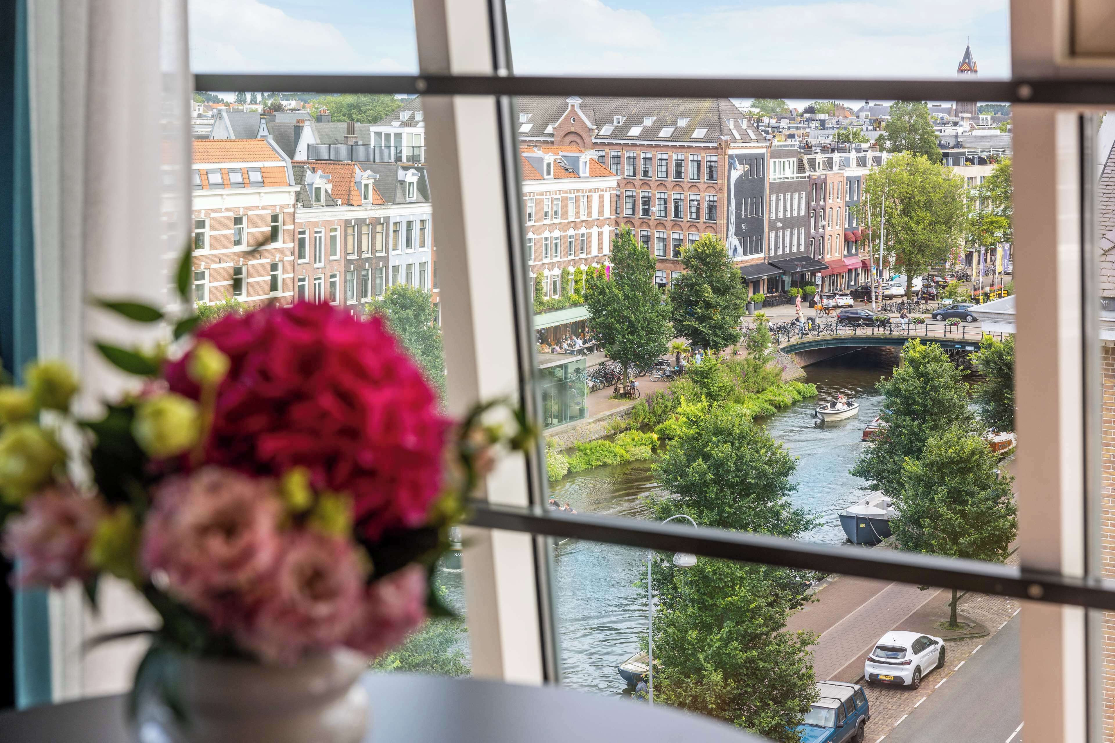 Blick durch ein Fenster auf einen Kanal mit Gebäuden, einer Brücke und einem Boot.