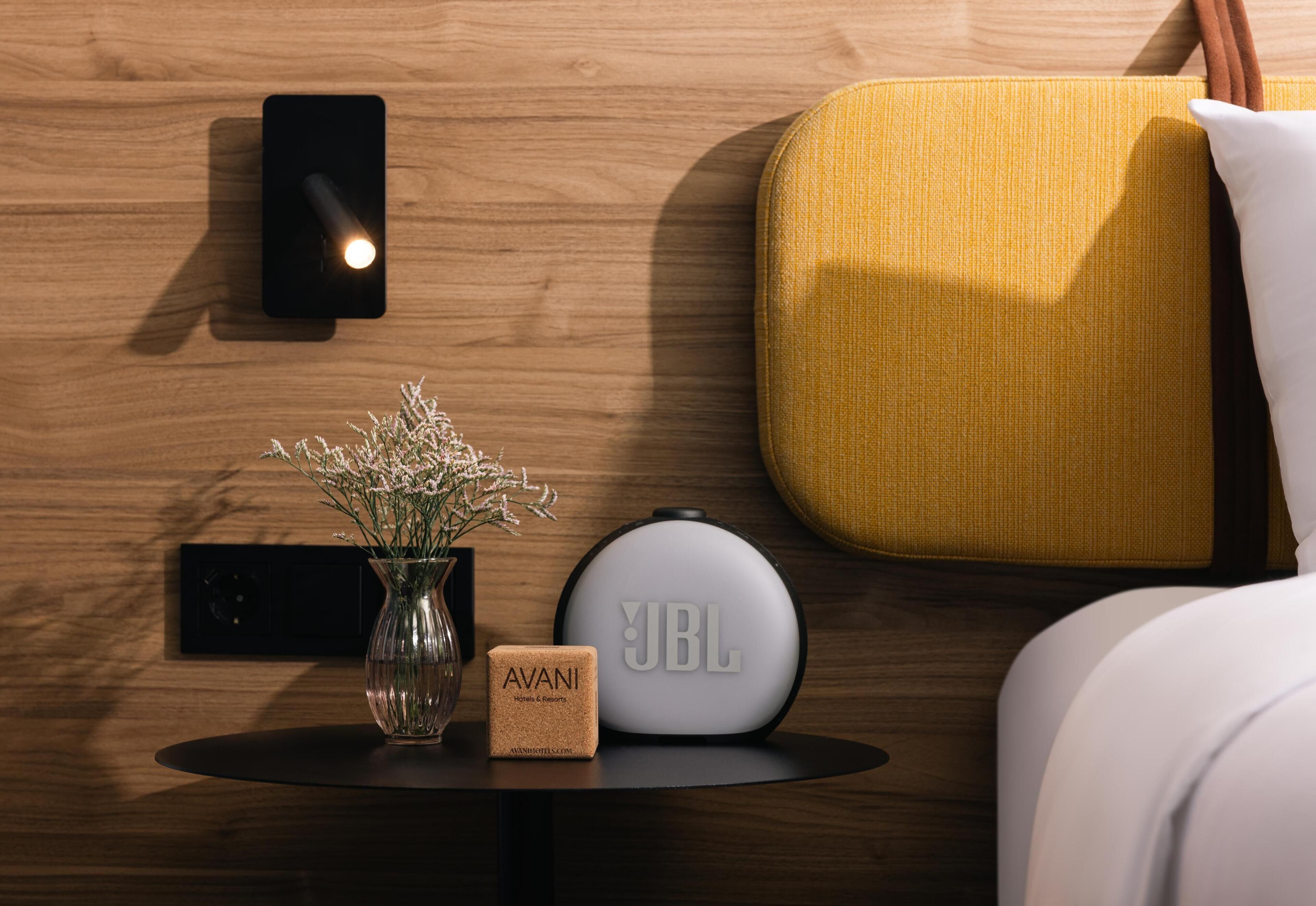 Habitación de hotel ordenada con pared de madera, un altavoz negro de pared, un altavoz blanco redondo, un pequeño jarrón con flores y una almohada amarilla sobre una cama.