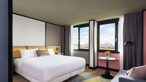 Une chambre d’hôtel avec un grand lit, une petite table sur laquelle est posé un vase, un canapé et une vue sur les immeubles par les fenêtres.