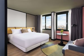 A hotel room with a large bed, a small table with a vase, a sofa, and a view of buildings through the window. - Avani Museum Quarter Amsterdam Hotel (Amsterdam)