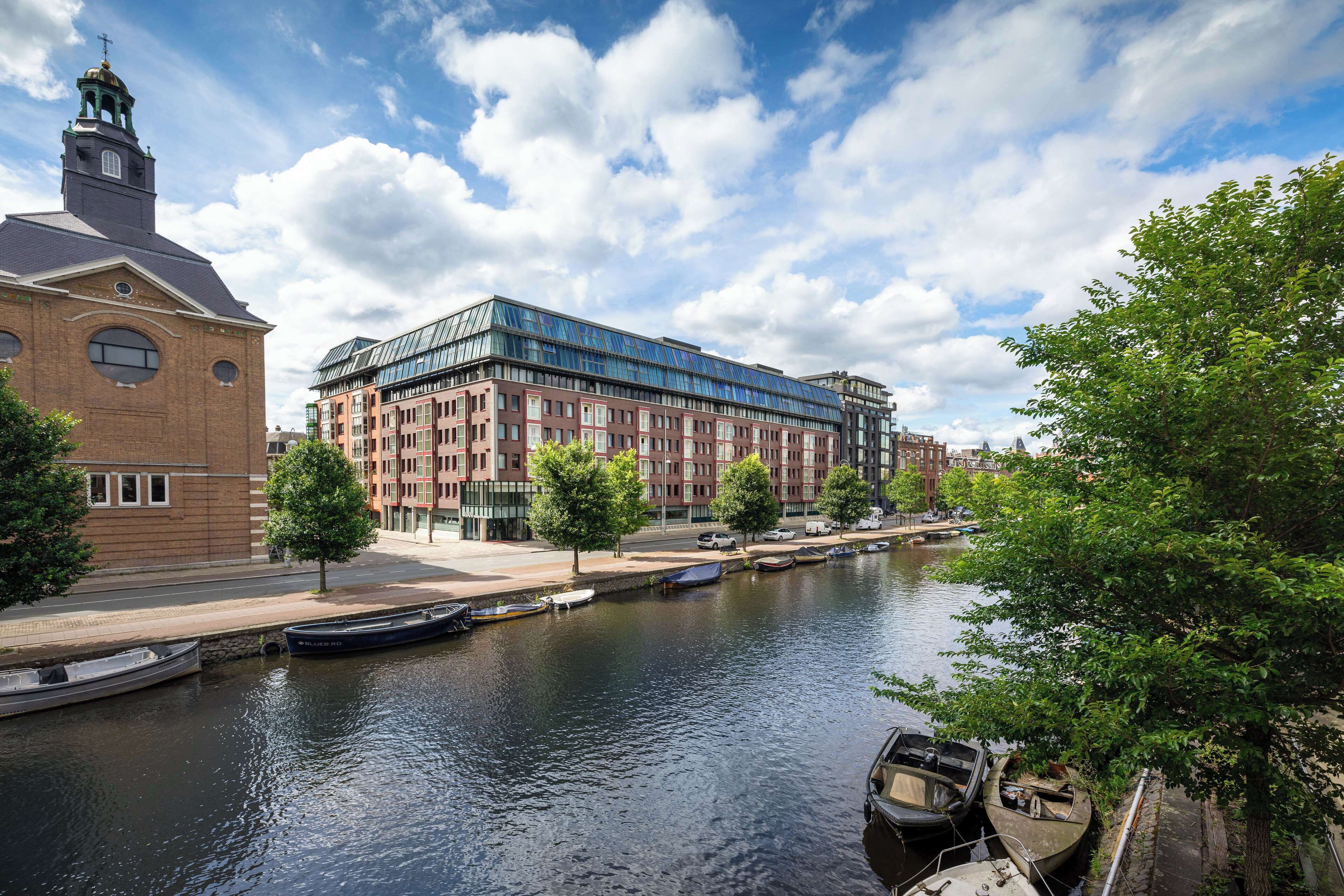 Ein Fluss mit vertäuten Booten rechts, ein modernes Gebäude mit Glasfassade und ein traditionelles Backsteingebäude mit einem Turm links.