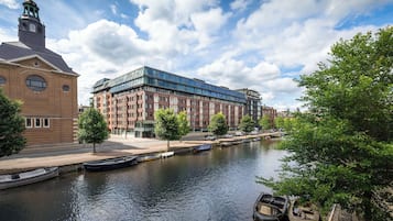 Ein Fluss mit vertäuten Booten rechts, ein modernes Gebäude mit Glasfassade und ein traditionelles Backsteingebäude mit einem Turm links.