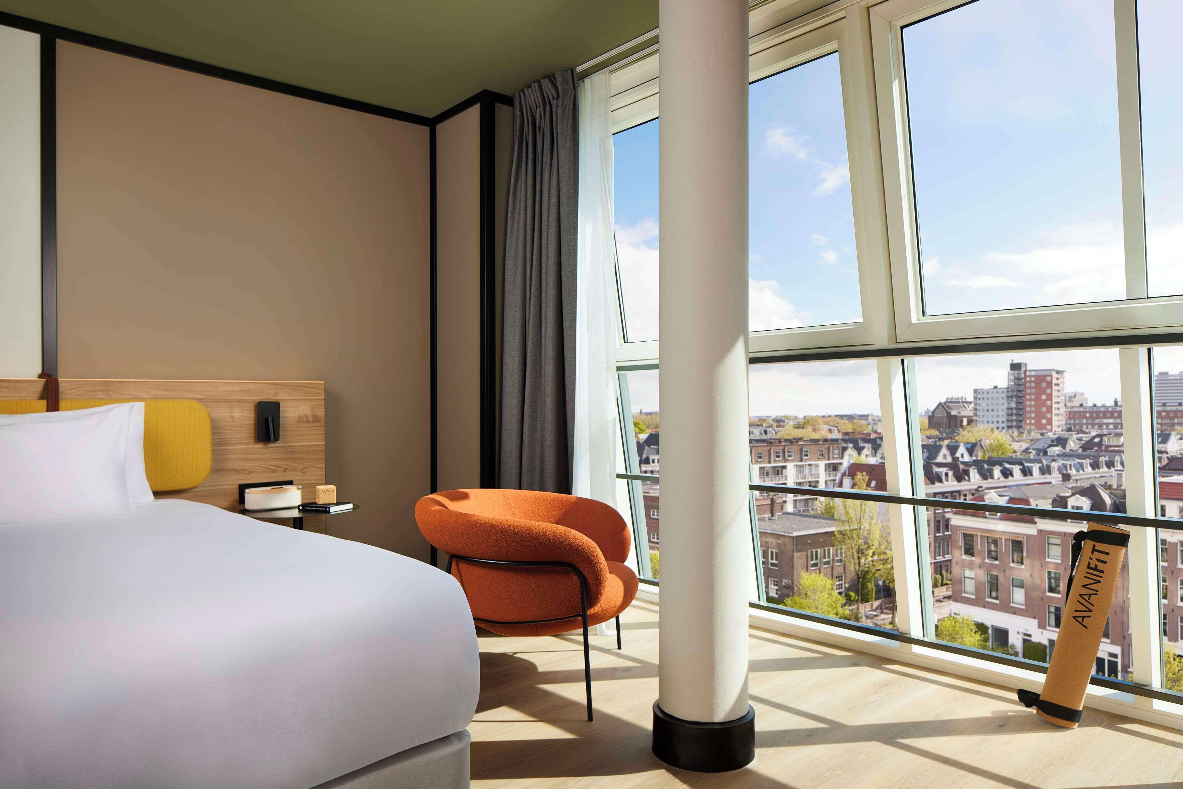 Quarto de hotel com uma grande janela, cama, uma cadeira e vista para a cidade.