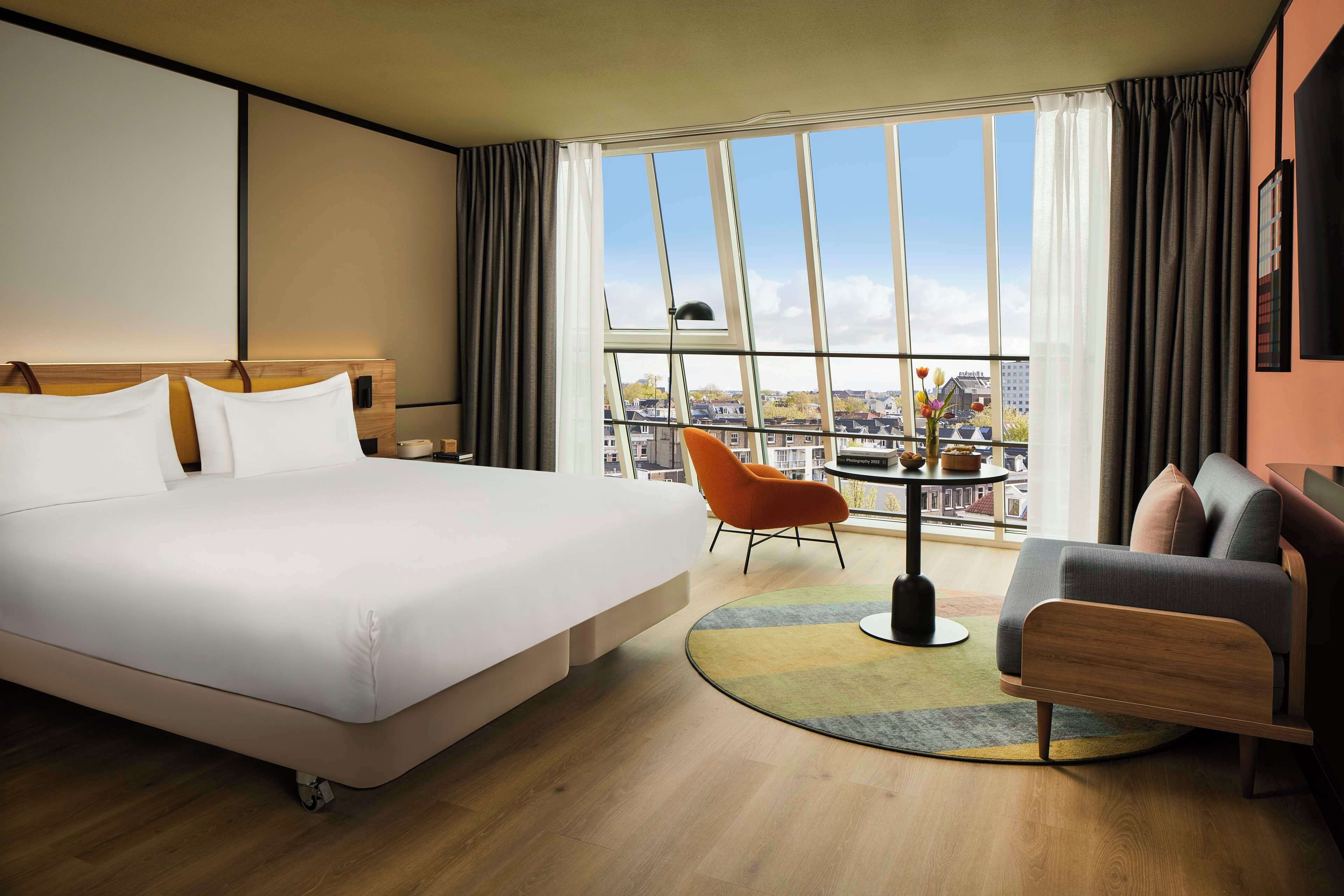 A hotel room with a large bed, a sofa, a small table, and a view of the city through large windows.
