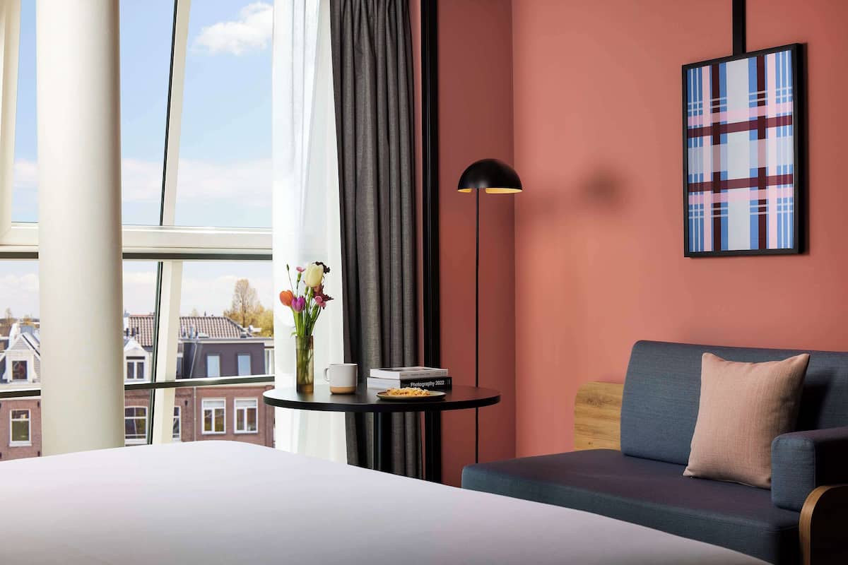 Ein Hotelzimmer mit Bett, Sofa, kleinem Tisch mit Vase und Büchern, Stehlampe und Fenster mit Blick auf Gebäude.