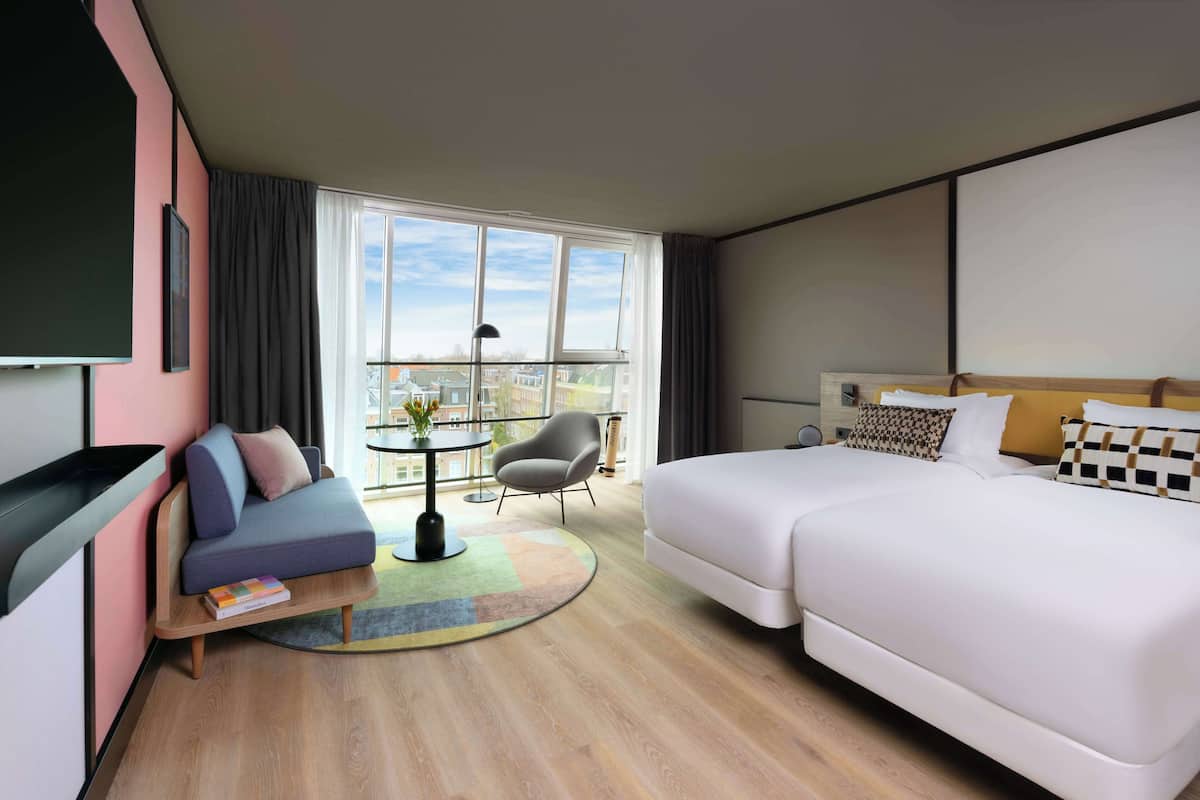 Una habitación de hotel moderna con sofá, cama, una mesita con un florero y vistas a la ciudad a través de amplios ventanales.