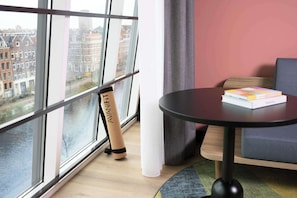 A modern room with a large window, a standing fan, a round table with books, and a view of buildings outside. - Avani Museum Quarter Amsterdam Hotel (Amsterdam)