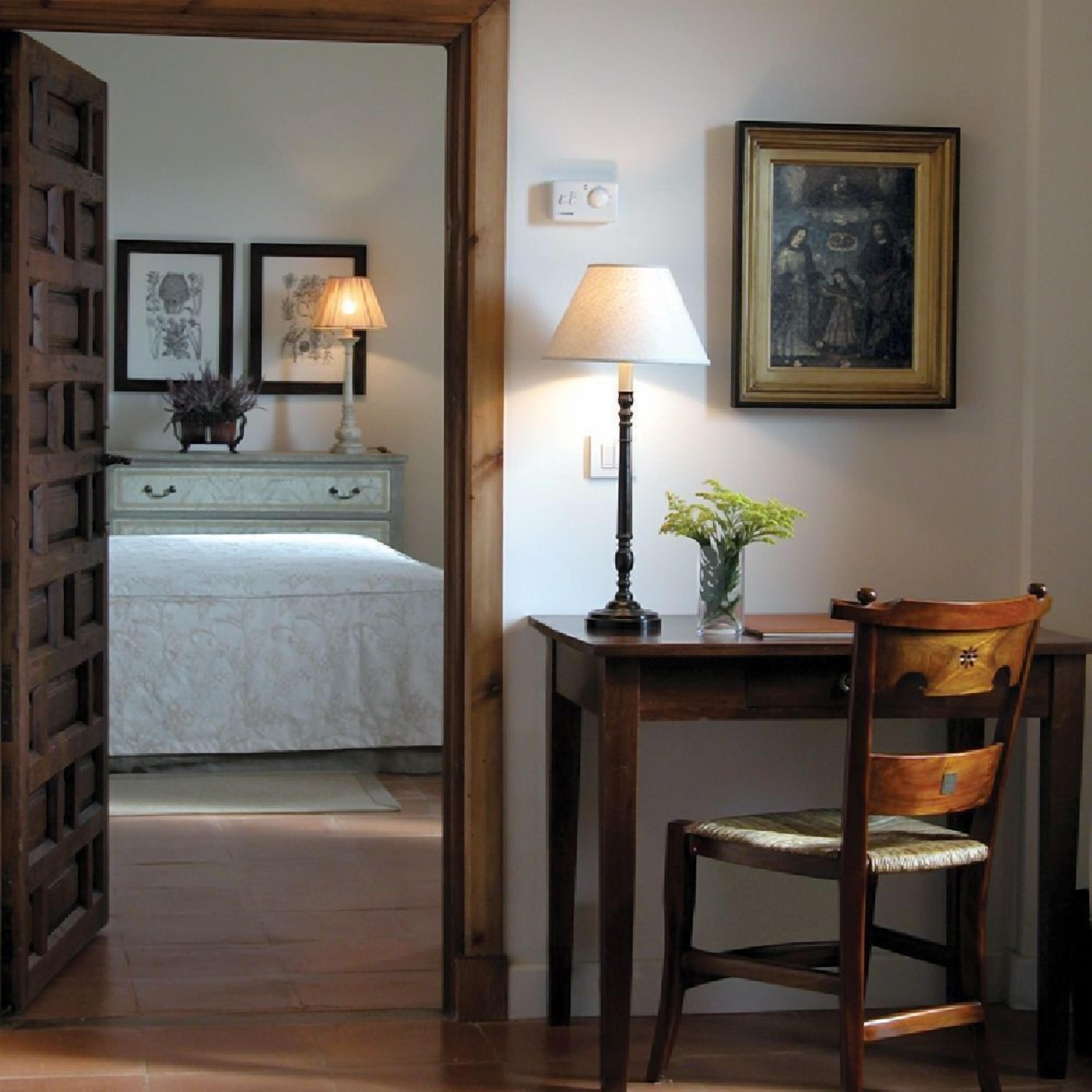 A bedroom with a bed, a nightstand with a lamp, a chair, and a desk with a plant.