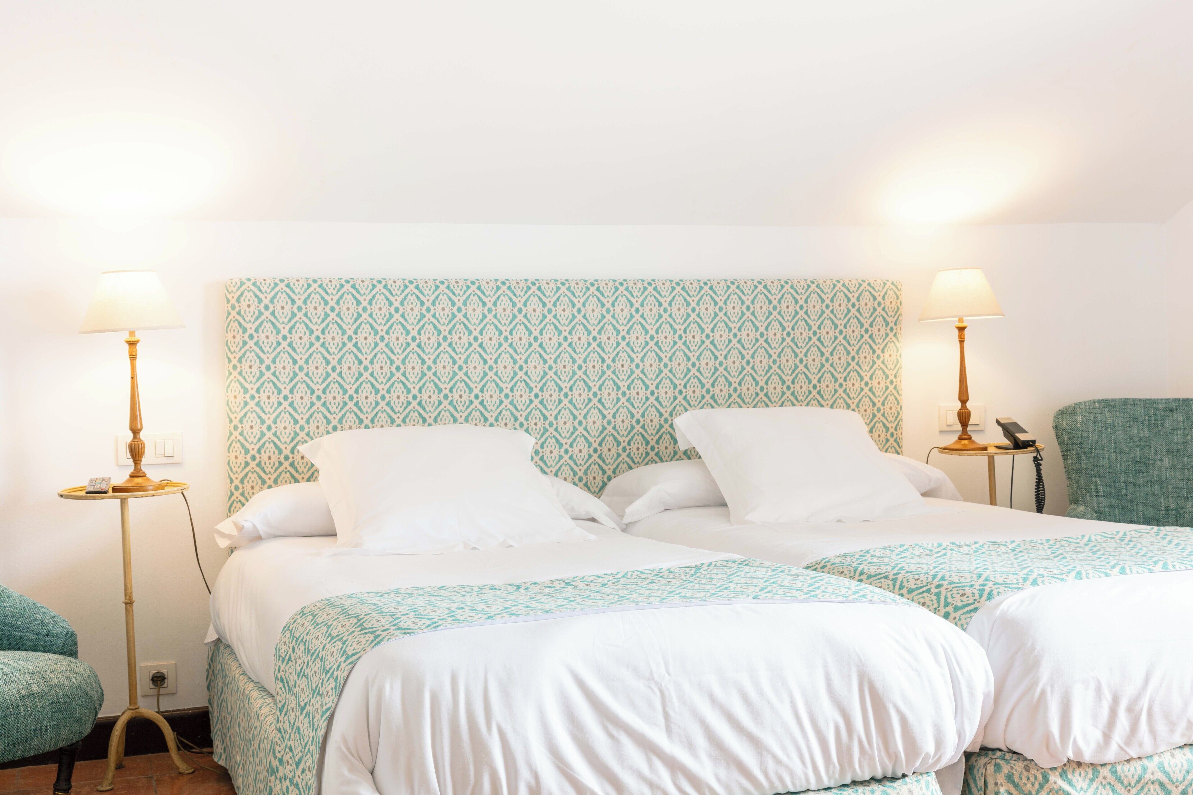 Quarto de hotel com duas camas, cada uma com cabeceira estampada, roupa de cama branca e duas luminárias laterais.