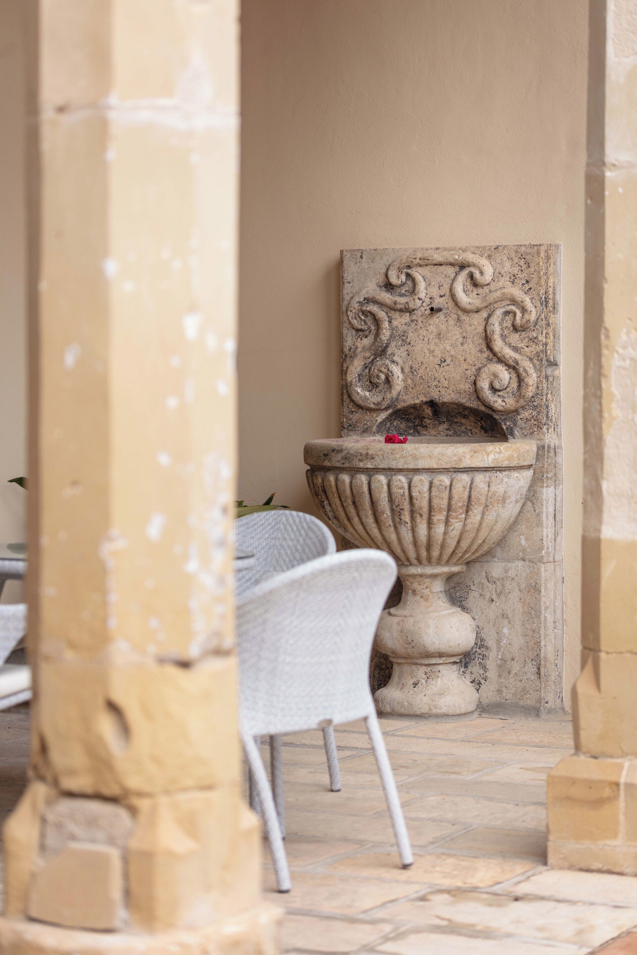 A stone fountain with intricate carvings, a white chair, and a tiled floor in a courtyard.