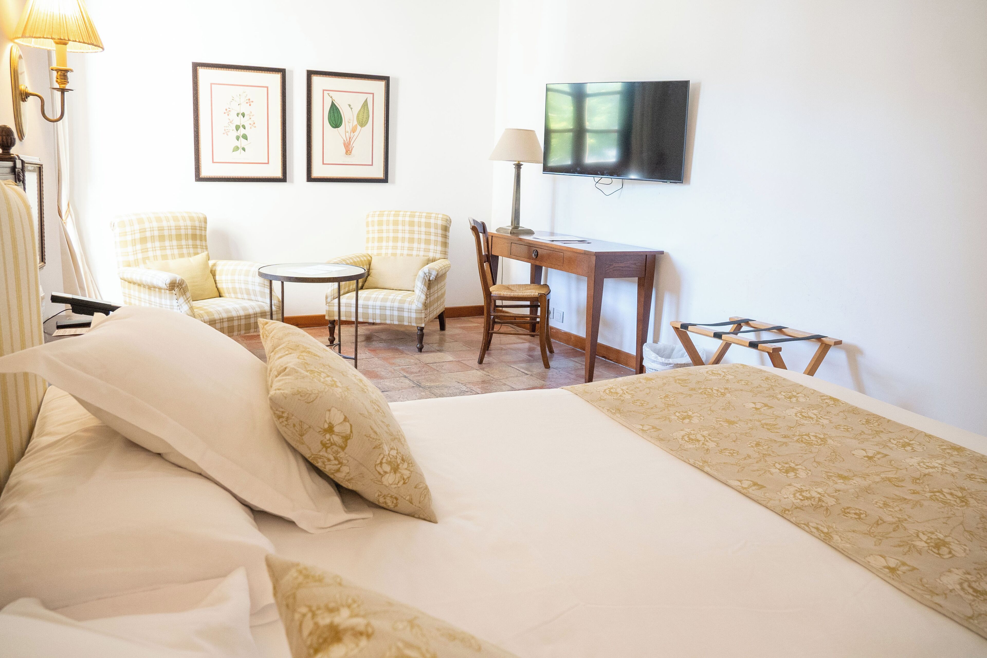 A hotel room with a bed, two chairs, a desk, a television, and framed pictures on the wall.