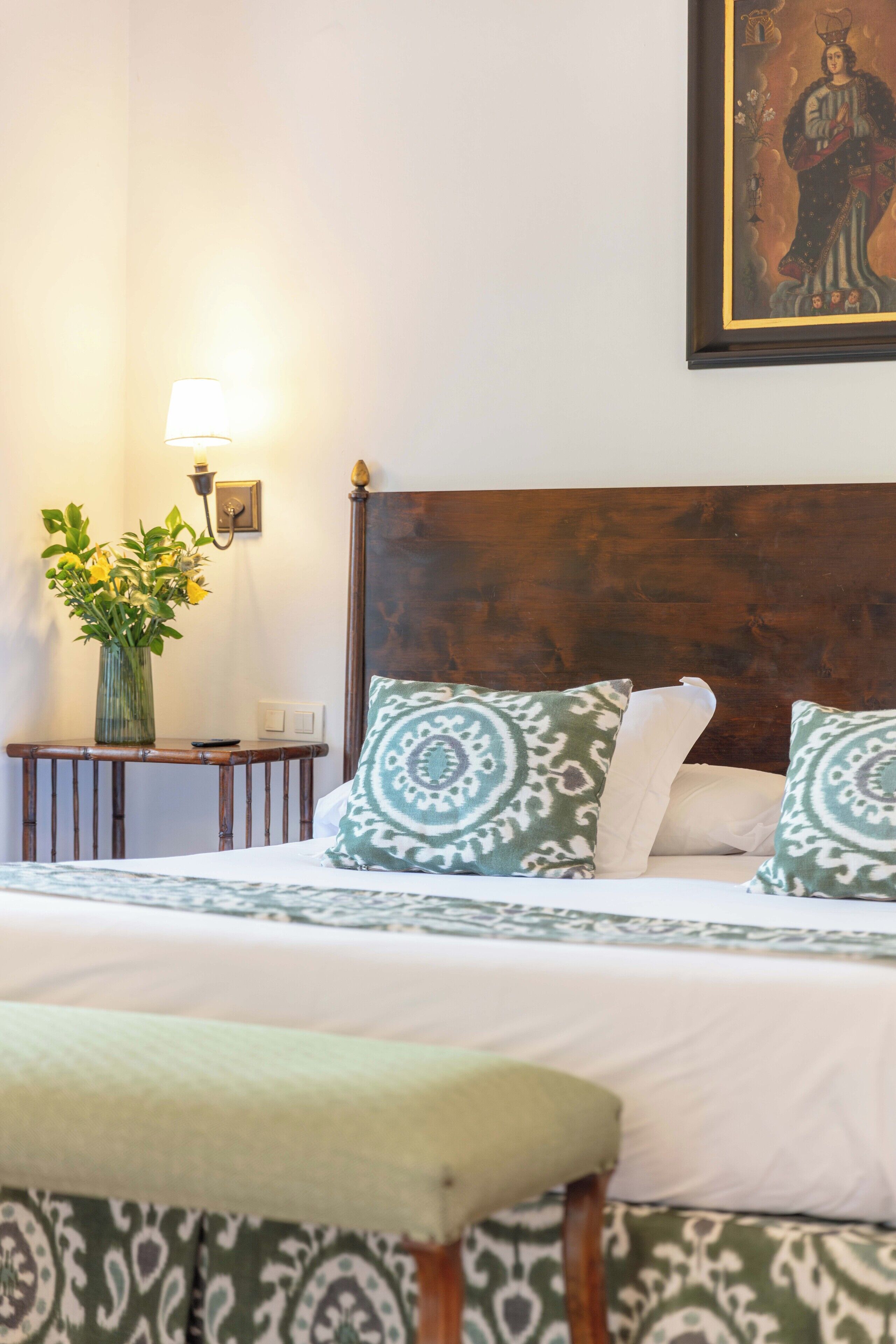 Un lit bien fait, agrémenté d’oreillers à motifs, une table de chevet avec un vase de fleurs et une lampe fixée au mur.
