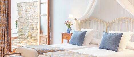 A bedroom with two beds, a stone wall, a wooden headboard, and a view of a stone building.