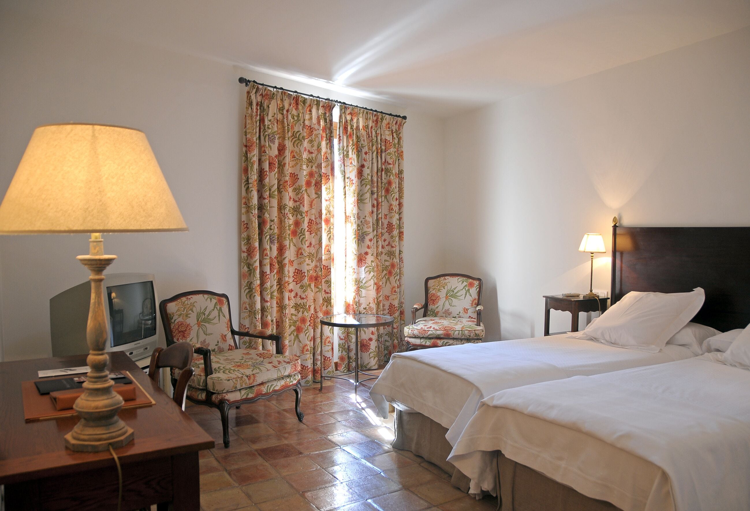 Habitación de hotel con dos camas, un escritorio con lámpara, una silla, una mesita y cortinas.