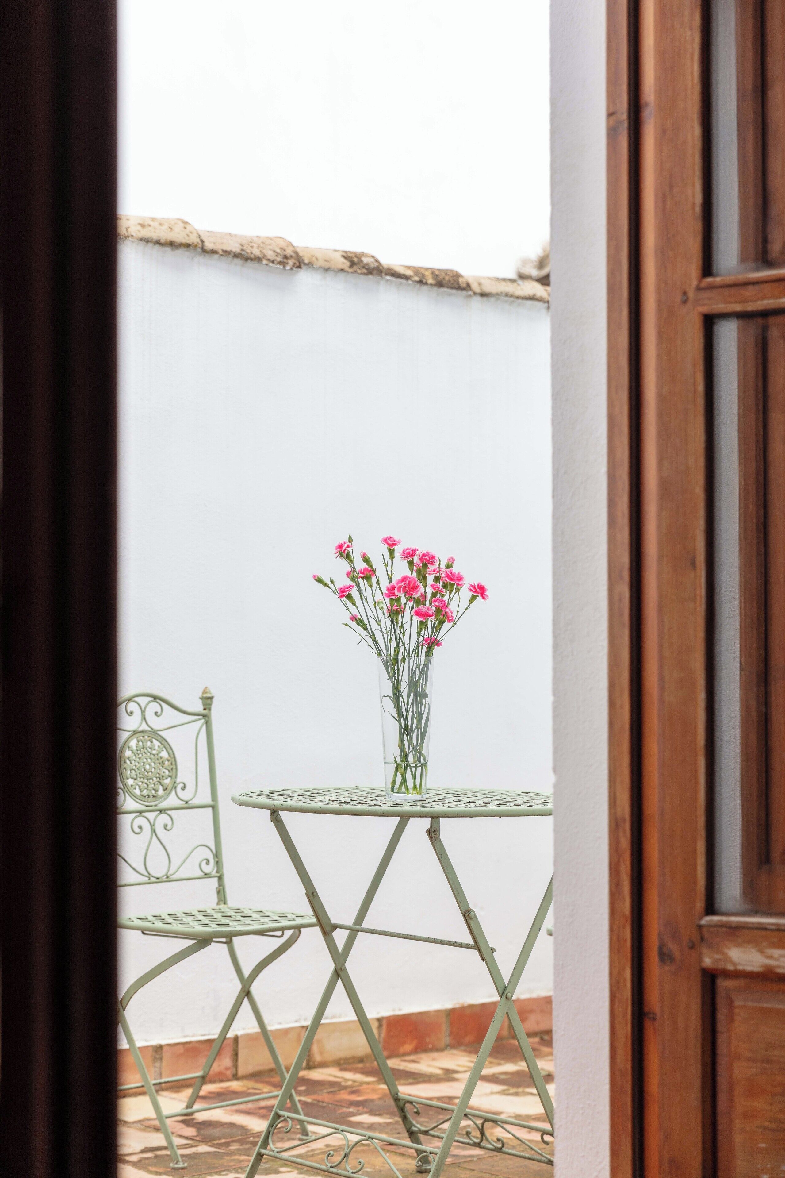 Une vue d’une terrasse avec une table et des chaises en métal, un vase de fleurs roses et un mur blanc en arrière-plan.