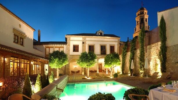 A courtyard with a swimming pool, outdoor seating, and a building with a bell tower. - Hotel Puerta de la Luna (Baeza)