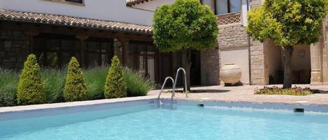 A swimming pool with a clear blue water, surrounded by a stone building with a tiled roof and a garden with trimmed bushes and trees.