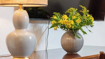 A wooden desk with a lamp, a vase with yellow flowers, and a television in the background.