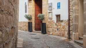 A narrow cobblestone street with a stone building featuring a large wooden door and barred windows. - Hotel Puerta de la Luna (Baeza)