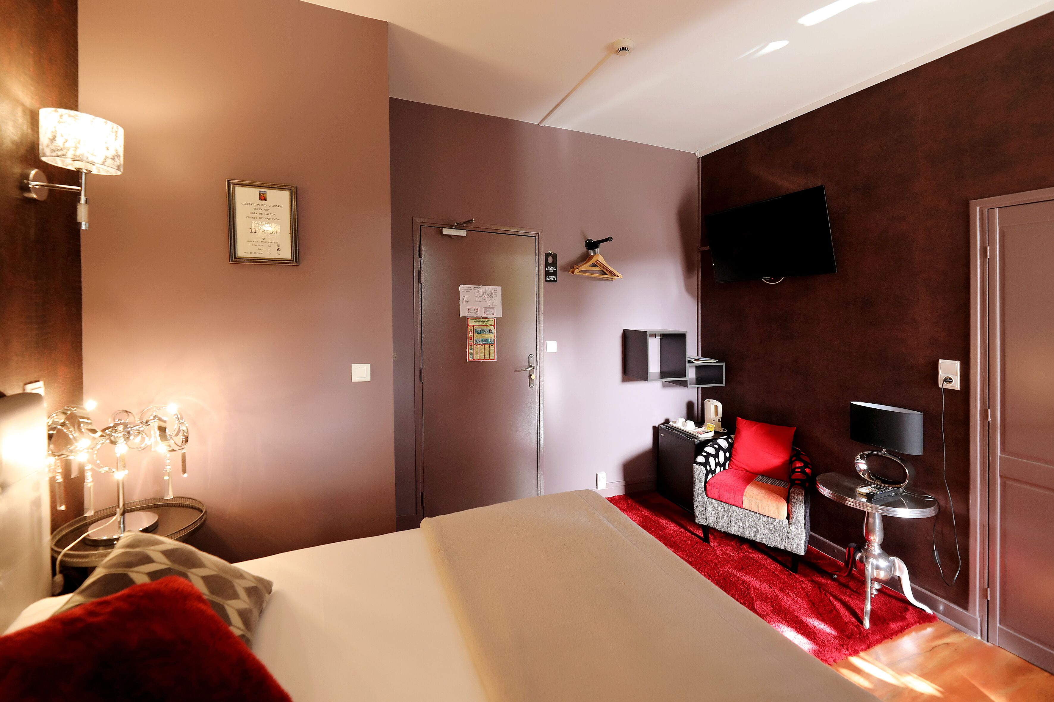 A hotel room with a bed, a red carpet, a TV, a small table, and a wall-mounted lamp.