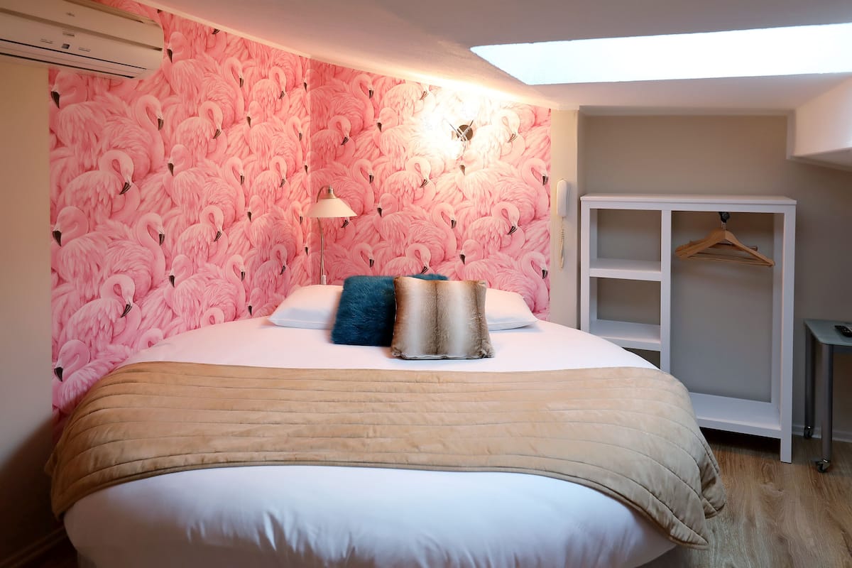A bedroom with a pink flamingo wallpaper, a bed with a beige blanket, a blue pillow, and a white bedside table.