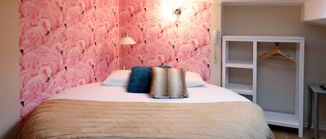 Un dormitorio con papel tapiz de flamencos rosas, una cama con una manta beige, un almohón azul y una mesita de noche blanca.