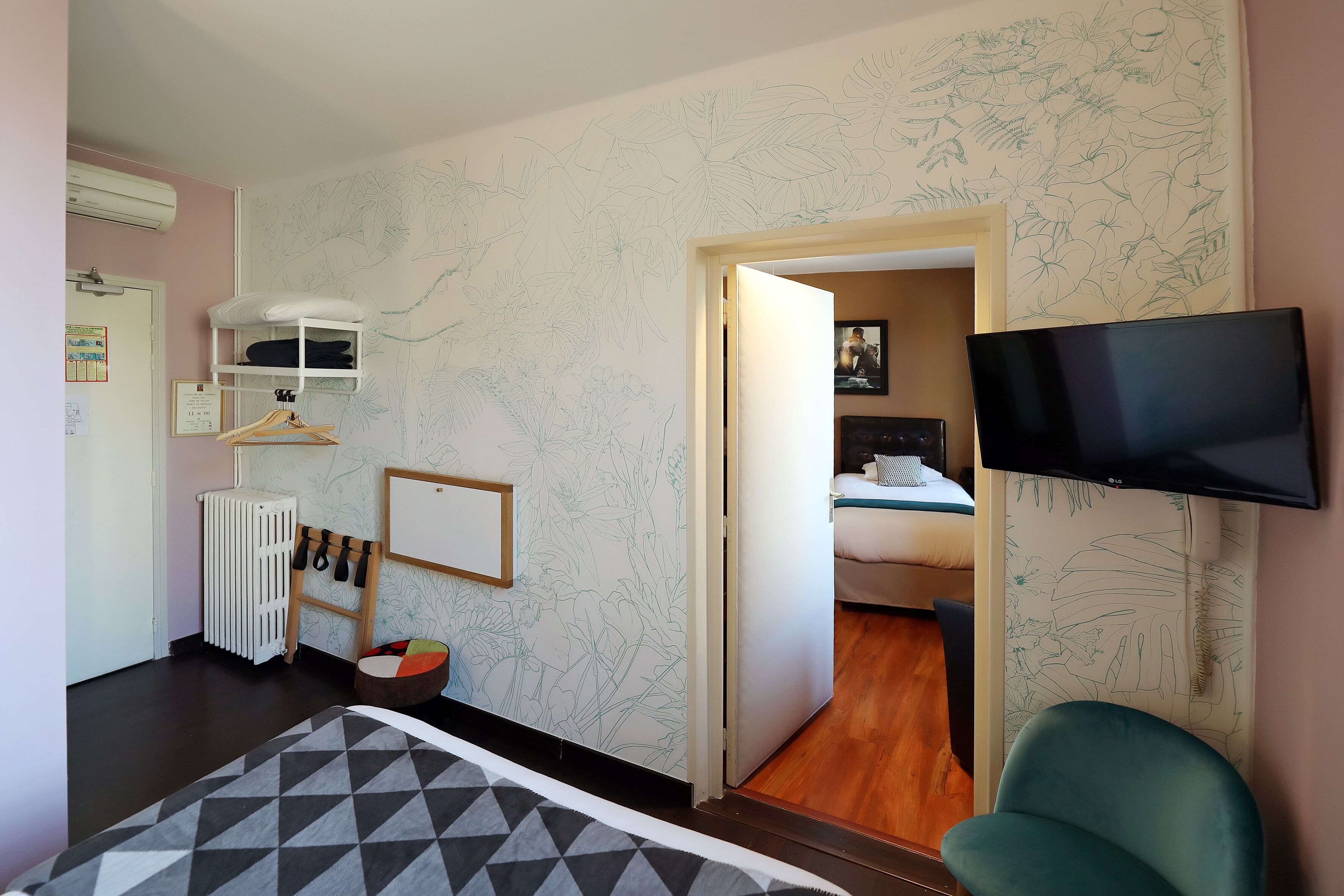 A room with a bed, a TV mounted on the wall, a radiator, a shelf with clothes, and a door leading to another room.