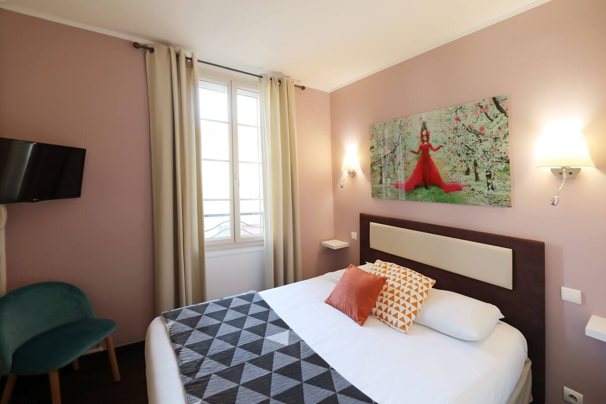 A hotel room with a bed, a television, a painting, and a window with curtains.