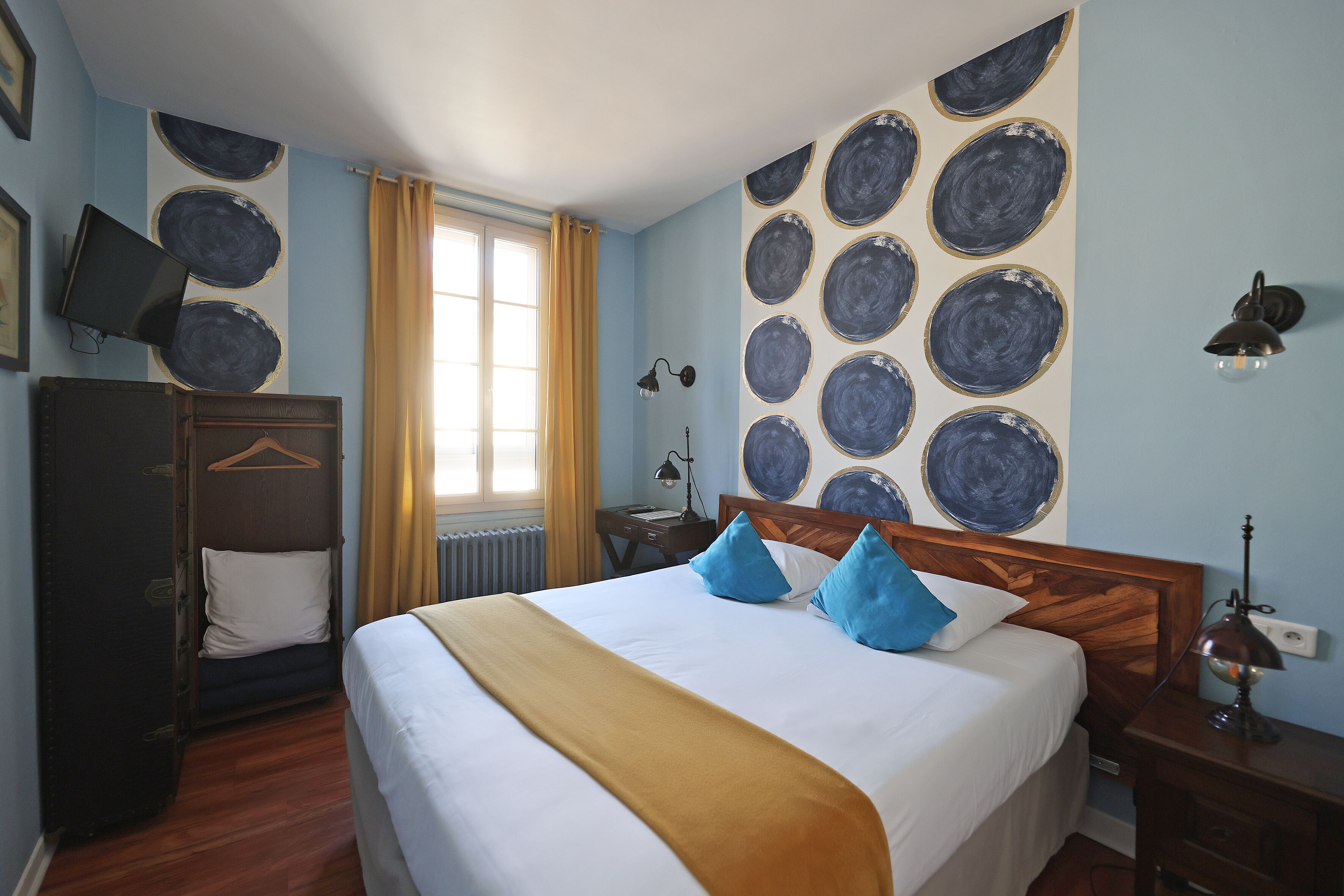 A hotel room with a large bed, a wooden wardrobe, a desk with a lamp, and a wall with circular patterns.